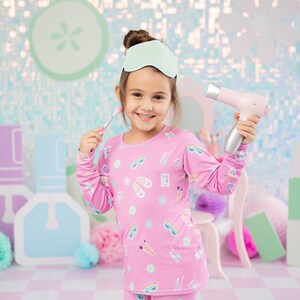 May include: A young girl wearing pink pajamas with a beauty-themed print is holding a pink hair dryer and a makeup brush. She is smiling and looking at the camera. The background is a light blue and pink sparkly wall.