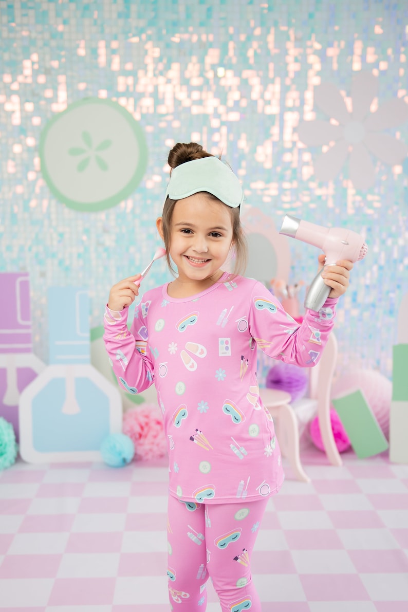 May include: A young girl wearing pink pajamas with a beauty-themed print is holding a pink hair dryer and a makeup brush. She is smiling and looking at the camera. The background is a light blue and pink sparkly wall.