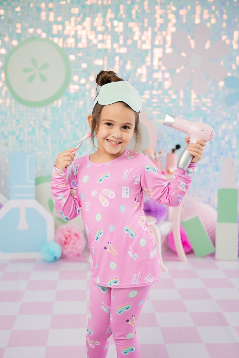 May include: A young girl wearing a pink long-sleeved pajama top and matching pants with a print of eye masks, makeup brushes, and other beauty items. She is holding a pink hairdryer and a makeup brush.