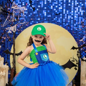 Puede incluir: Un ni&ntilde;o vestido como un personaje de videojuego para Halloween. Lleva un disfraz verde y azul con un sombrero verde y un bigote falso. El disfraz incluye una falda de tul azul. El ni&ntilde;o est&aacute; de pie frente a un fondo azul y amarillo con murci&eacute;lagos y una luna.