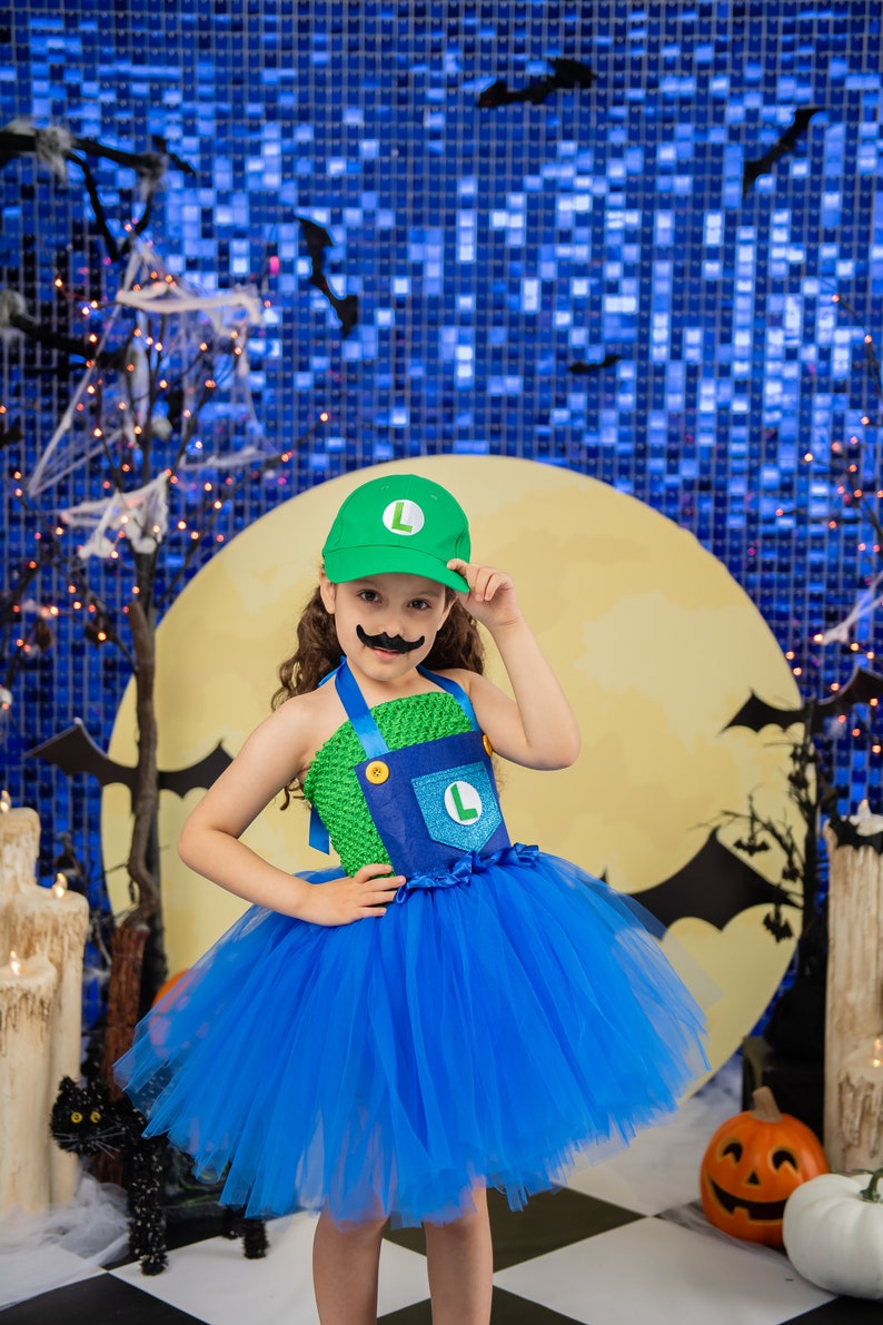 Puede incluir: Un ni&ntilde;o vestido como un personaje de videojuego para Halloween. Lleva un disfraz verde y azul con un sombrero verde y un bigote falso. El disfraz incluye una falda de tul azul. El ni&ntilde;o est&aacute; de pie frente a un fondo azul y amarillo con murci&eacute;lagos y una luna.