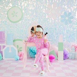 May include: A young girl wearing a pink pajama set with a sleep mask sits at a vanity table with a mirror and makeup brushes. The vanity table is pink and has a white stool. The background is a light blue and pink sparkly wall with paper flowers.