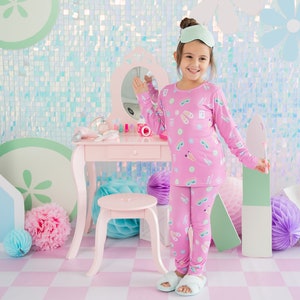 May include: A young girl wearing pink pajamas with a print of eye masks and sandals stands in front of a vanity set. The vanity is pink with a mirror and a drawer. The girl is smiling and holding a green and white object in her right hand.
