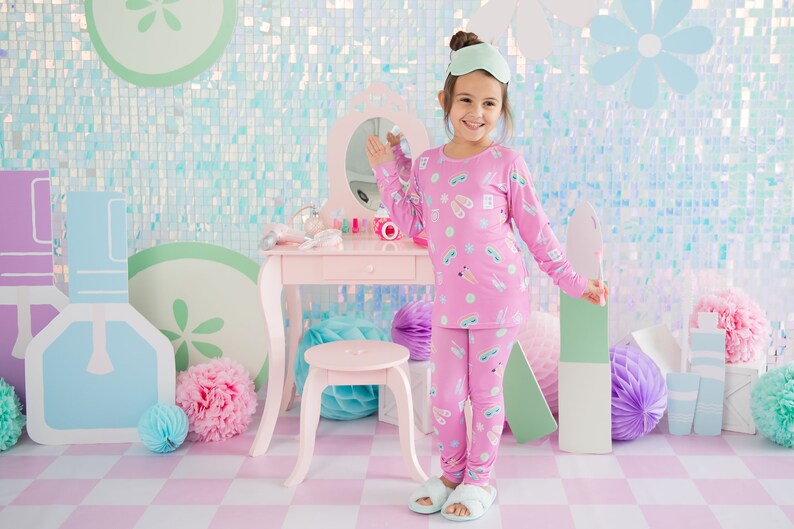 May include: A young girl wearing pink pajamas with a print of eye masks and sandals stands in front of a vanity set. The vanity is pink with a mirror and a drawer. The girl is smiling and holding a green and white object in her right hand.