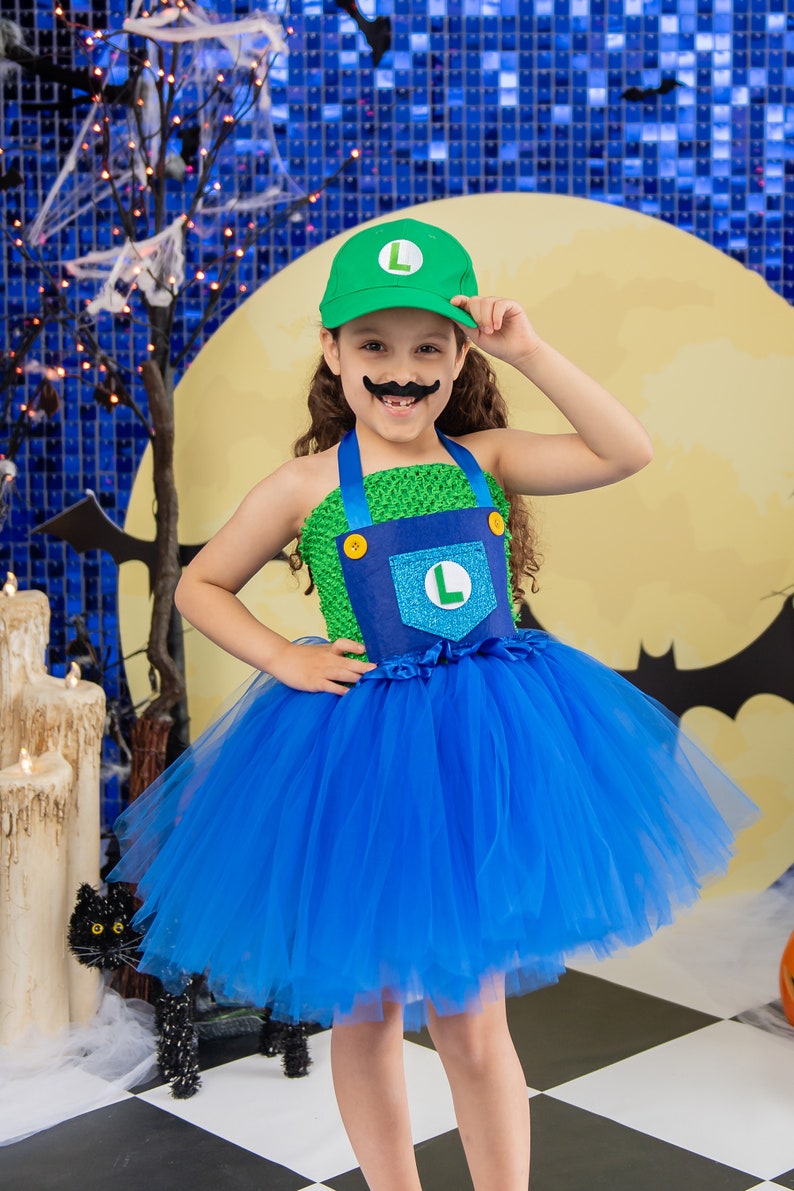 Puede incluir: Un ni&ntilde;o vestido como un personaje de videojuego para Halloween. Lleva un sombrero verde con una 'L' blanca en &eacute;l, un vestido verde y azul con una 'L' azul en el bolsillo y un bigote falso. El ni&ntilde;o est&aacute; de pie sobre un suelo de ajedrez negro y blanco.