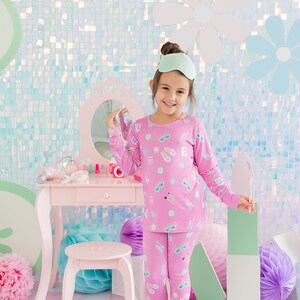 May include: A young girl wearing pink pajamas with a sleep mask on her head. The pajamas have a pattern of various spa items, including eye masks, slippers, and bath bombs. The girl is standing in front of a vanity table with a mirror and makeup supplies.