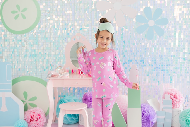 May include: A young girl wearing pink pajamas with a sleep mask on her head. The pajamas have a pattern of various spa items, including eye masks, slippers, and bath bombs. The girl is standing in front of a vanity table with a mirror and makeup supplies.
