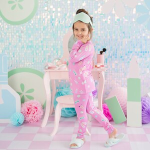 May include: A young girl wearing pink pajamas with a makeup theme print. She is standing in front of a vanity set with a mirror and a stool. The background is a light blue and green iridescent wall with cutouts of makeup bottles and flowers.
