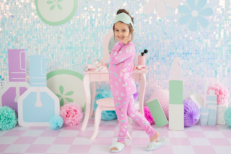 May include: A young girl wearing pink pajamas with a makeup theme print. She is standing in front of a vanity set with a mirror and a stool. The background is a light blue and green iridescent wall with cutouts of makeup bottles and flowers.
