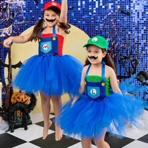 Puede incluir: Dos ni&ntilde;os disfrazados de Mario y Luigi para Halloween. Llevan tut&uacute;s azules y monos con los ic&oacute;nicos s&iacute;mbolos 'M' y 'L'. La ni&ntilde;a disfrazada de Mario tiene el brazo levantado en el aire, mientras que la ni&ntilde;a disfrazada de Luigi sonr&iacute;e. Est&aacute;n de pie sobre un suelo de baldosas blancas y negras.