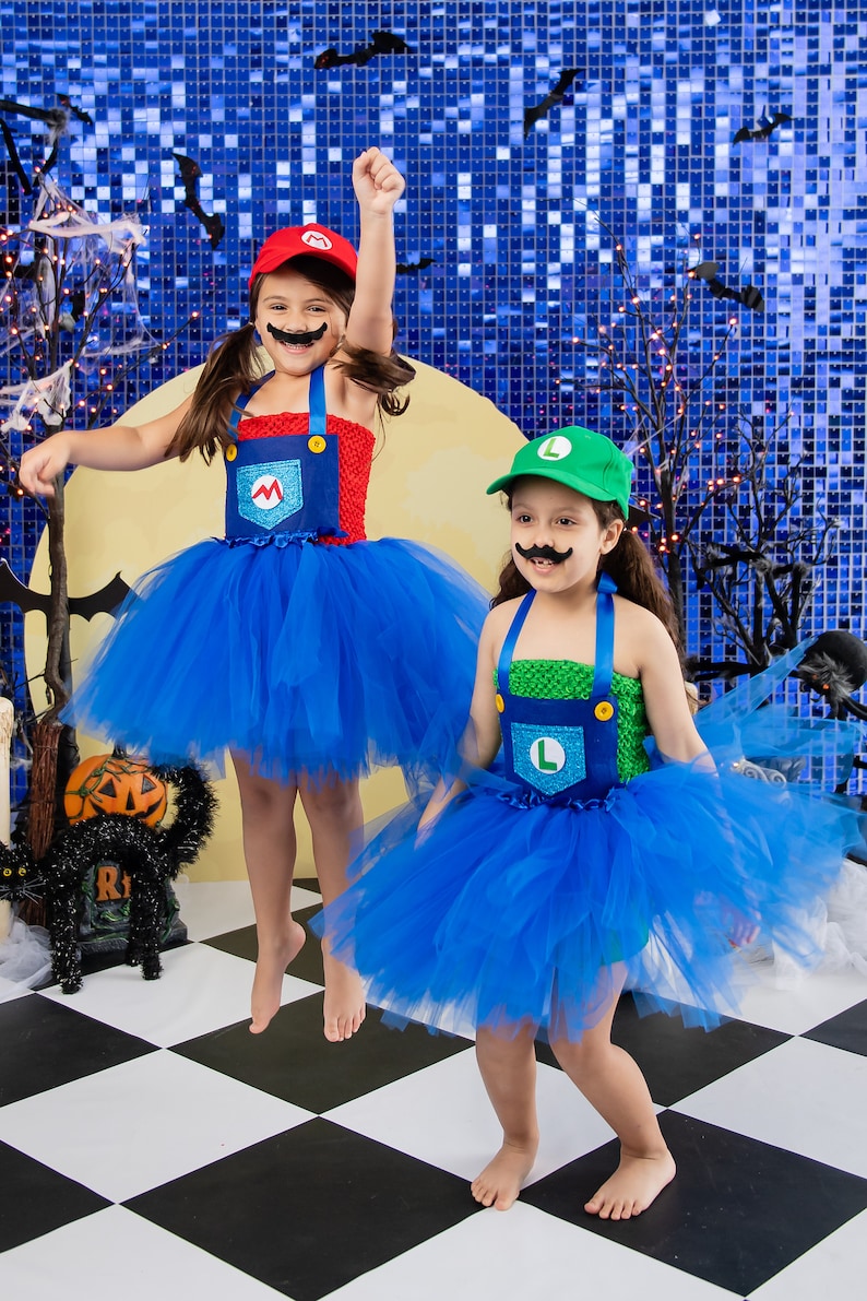Puede incluir: Dos ni&ntilde;os disfrazados de Mario y Luigi para Halloween. Llevan tut&uacute;s azules y monos con los ic&oacute;nicos s&iacute;mbolos 'M' y 'L'. La ni&ntilde;a disfrazada de Mario tiene el brazo levantado en el aire, mientras que la ni&ntilde;a disfrazada de Luigi sonr&iacute;e. Est&aacute;n de pie sobre un suelo de baldosas blancas y negras.