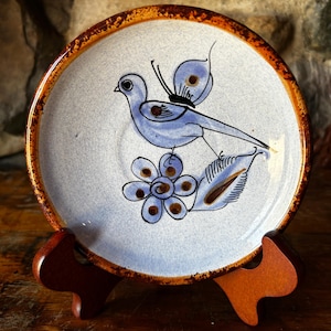 Puede incluir: Un platillo de cerámica redondo con esmalte moteado azul claro y borde marrón oscuro. El centro presenta un diseño pintado a mano de un pájaro azul con una mariposa en la espalda, posado sobre una flor azul y una hoja con detalles marrones.