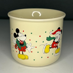 Peut inclure: Mug en céramique crème avec des illustrations de Mickey Mouse festives. Un Mickey porte une couronne, l'autre patine. Le mug est décoré de pois rouges et verts, créant un thème de Noël joyeux.