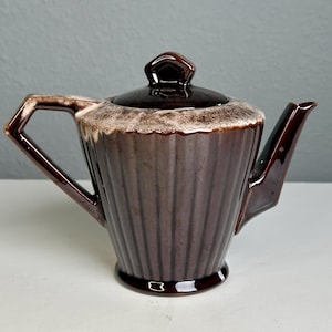 Könnte beinhalten: Eine braune Keramik-Teekanne mit Deckel. Die Teekanne hat ein geripptes Design und einen Griff an der Seite. Der Rand der Teekanne hat eine hellbraune, strukturierte Glasur. Der Ausguss ist eckig. Die Teekanne steht auf einer weißen Oberfläche.