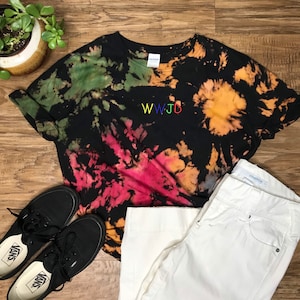 Puede incluir: Una camiseta negra tie-dye con las palabras "WWJD" bordadas en colores arcoíris en la parte delantera. La camiseta se combina con jeans blancos y zapatos Vans negros.