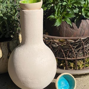 Könnte beinhalten: Eine handgefertigte, cremefarbene Keramikvase mit rundem Boden und einer kleinen, grün gefütterten Schale oben. Eine zweite Schale mit leuchtend blauem Innenraum befindet sich in der Nähe. Die Vase wird im Freien mit Grün im Hintergrund ausgestellt.