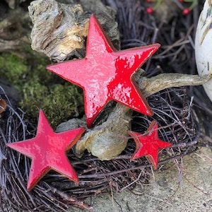 Op de afbeelding: Drie glanzende rode sterornamenten van verschillende grootte rusten op een bed van twijgen, mos en verweerd hout. De sterren hebben een rustieke, handgemaakte uitstraling, perfect voor vakantie- of seizoensdecoratie. De opstelling staat tegen een natuurlijke achtergrond.