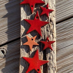 Op de afbeelding: Een verzameling rode, stervormige ornamenten met een glanzende afwerking, gerangschikt op een verweerde houten plank. De sterren variëren in grootte en hebben kleine gaatjes om op te hangen. Het hout heeft een natuurlijke, rustieke uitstraling.