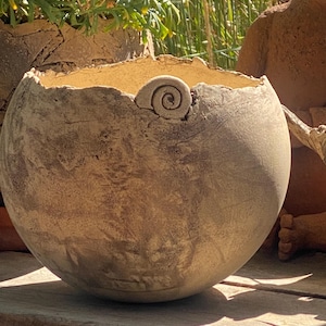 Può includere: Una ciotola in ceramica beige di forma rotonda con un bordo ruvido e irregolare e una superficie strutturata e maculata. Un piccolo dettaglio in argilla a spirale è fissato sul bordo superiore della ciotola, che presenta un'ampia apertura.