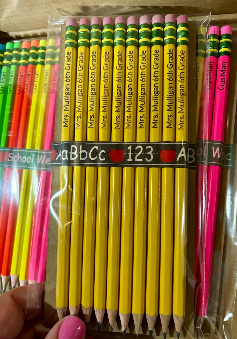 May include: A close-up of a package of yellow pencils with green and pink eraser tops. The pencils are labeled "Mrs. Mulligan 6th Grade". There are also some pink and yellow pencils in the background.