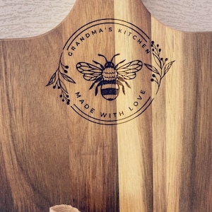 May include: A wooden cutting board with a circular design featuring a bee and the text "Grandma's Kitchen Made With Love".