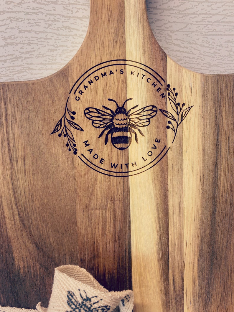 May include: A wooden cutting board with a circular design featuring a bee and the text "Grandma's Kitchen Made With Love".