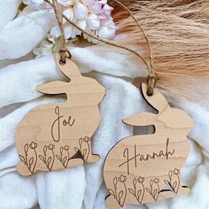 Puede incluir: Dos adornos de conejo de madera con detalles florales y nombres personalizados, "Joe" y "Hannah", colgados de un cordel.