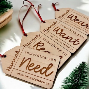 Puede incluir: Cuatro etiquetas de regalo de madera con cinta roja, cada una con el nombre "Hannah" y el texto "something you", seguido de "wear", "want", "read" y "need". Cada etiqueta también tiene el texto "OPEN ON DECEMBER 25".