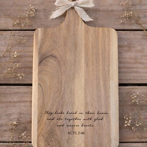 Puede incluir: Tabla de cortar de madera con asa y lazo claro. La tabla tiene grabado el texto "They broke bread in their homes and ate together with glad and sincere hearts. ACTS 2:46". La tabla est&aacute; sobre una superficie de madera.