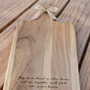 Puede incluir: Tabla de cortar de madera con asa y lazo decorativo. La tabla presenta texto grabado de Hechos 2:46: "Part&iacute;an el pan por las casas y com&iacute;an juntos con alegr&iacute;a y sencillez de coraz&oacute;n."