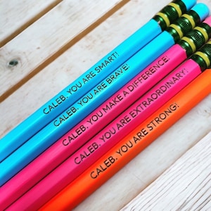 Puede incluir: Cuatro lápices de colores con mensajes alentadores para una persona llamada Caleb. Los lápices son azul, rosa, naranja y rojo. Los mensajes en los lápices son "Caleb. ¡Eres inteligente!", "Caleb. ¡Eres valiente!", "Caleb. ¡Haces la diferencia!", "Caleb. ¡Eres extraordinario!" y "Caleb. ¡Eres fuerte!"