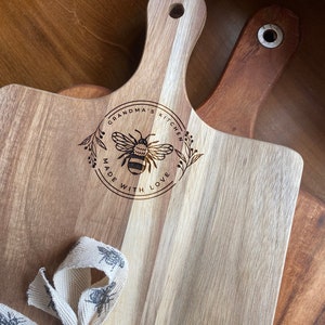 May include: A wooden cutting board with a handle, featuring a bee design and the text "Grandma's Kitchen Made With Love". The board is tied with a white ribbon with a bee pattern.