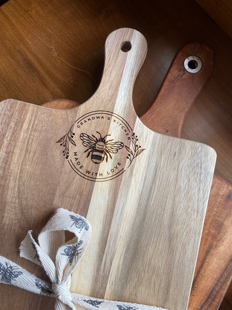 May include: A wooden cutting board with a handle, featuring a bee design and the text "Grandma's Kitchen Made With Love". The board is tied with a white ribbon with a bee pattern.