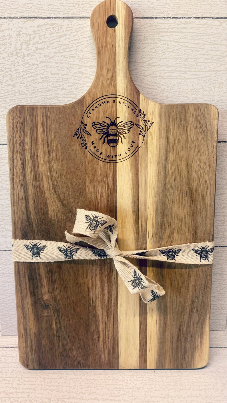 May include: A wooden cutting board with a handle, featuring a bee design and the text "Grandma's Kitchen Made With Love". The board is tied with a beige ribbon with a bee pattern.