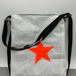 Könnte beinhalten: Weiße Canvas-Schultertasche mit einem leuchtend orangefarbenen Sternendesign und schwarzen verstellbaren Trägern.