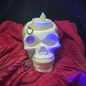 May include: A white skull-shaped candle holder with a flickering LED candle. A silver necklace with a cross pendant and a circular pendant hangs from the skull. The skull rests on a red, textured fabric, with a dark background.