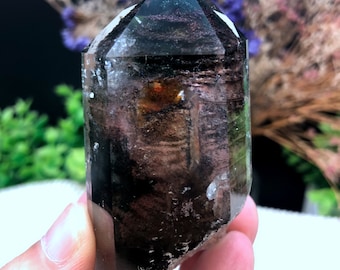 May include: A clear quartz crystal point with black and brown inclusions. The crystal is approximately 2 inches tall and has a pointed tip. The crystal is a natural formation and has a unique and beautiful appearance.