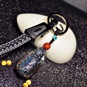 Puede incluir: Un llavero de cuero negro con una correa trenzada y un anillo de llave plateado. Un colgante de piedra iridiscente oscura con un diseño tallado cuelga del anillo. El colgante está unido al anillo con un cordón marrón y cuentas turquesas.