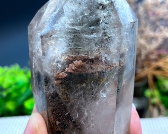 May include: A clear quartz crystal point with inclusions of brown and green minerals. The crystal is approximately 5 inches tall and has a pointed tip.