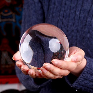 May include: A clear glass sphere, also known as a crystal ball, is held in the hands of a person. The sphere is reflecting the person's hand and the background.