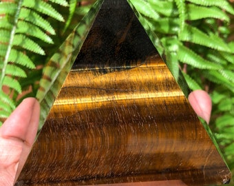 May include: A golden brown tiger's eye pyramid crystal with a dark brown top. The pyramid is sitting on a hand.