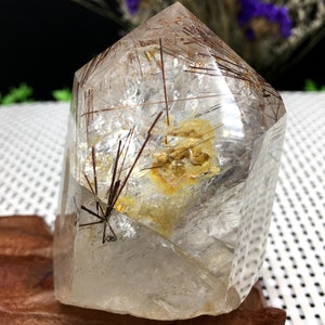 May include: A clear quartz crystal with red and gold inclusions. The crystal is shaped like a point and is sitting on a wooden base.
