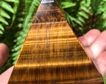 May include: A polished, golden brown tiger's eye pyramid crystal. The crystal has a striated pattern and a smooth, polished surface.