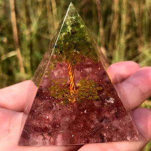 May include: A clear glass pyramid with a copper wire tree inside. The tree is surrounded by green and gold glitter. The base of the pyramid is filled with pink and white crystals.