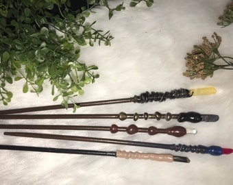 May include: Five wooden wands with different colored tips and decorative details. The wands are arranged in a row on a white surface.