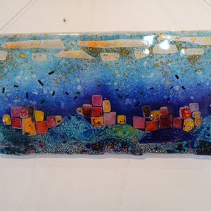 Op de afbeelding: Gesmolten glaskunstwerk dat een stadsgezicht toont met een blauwe lucht en kleurrijke gebouwen. Het kunstwerk heeft een gelaagd ontwerp met verschillende tinten blauw, oranje, geel en roze. Het rechthoekige stuk hangt aan een muur.
