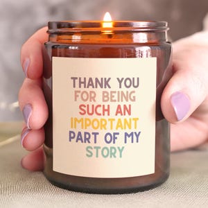 May include: A burning candle in a brown glass jar with a cream label. The label displays the text "THANK YOU FOR BEING SUCH AN IMPORTANT PART OF MY STORY" in multiple colours. The candle is held by a person.