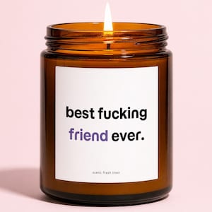 May include: A lit candle in a brown glass jar with a white label. The label reads "best fucking friend ever" in black and purple text. The scent is "fresh linen".