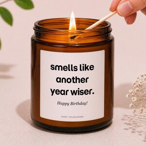 May include: A lit brown glass candle jar with a white label that reads "smells like another year wiser. Happy Birthday!" The candle is being lit with a match. The scent is wildflowers.
