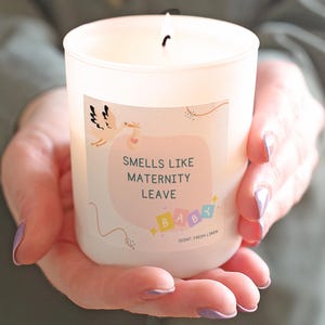 May include: A burning white candle in a glass jar, labelled "Smells Like Maternity Leave." The label shows a stork with a baby and the word "Baby" in colourful letters. The scent is "Fresh Linen."